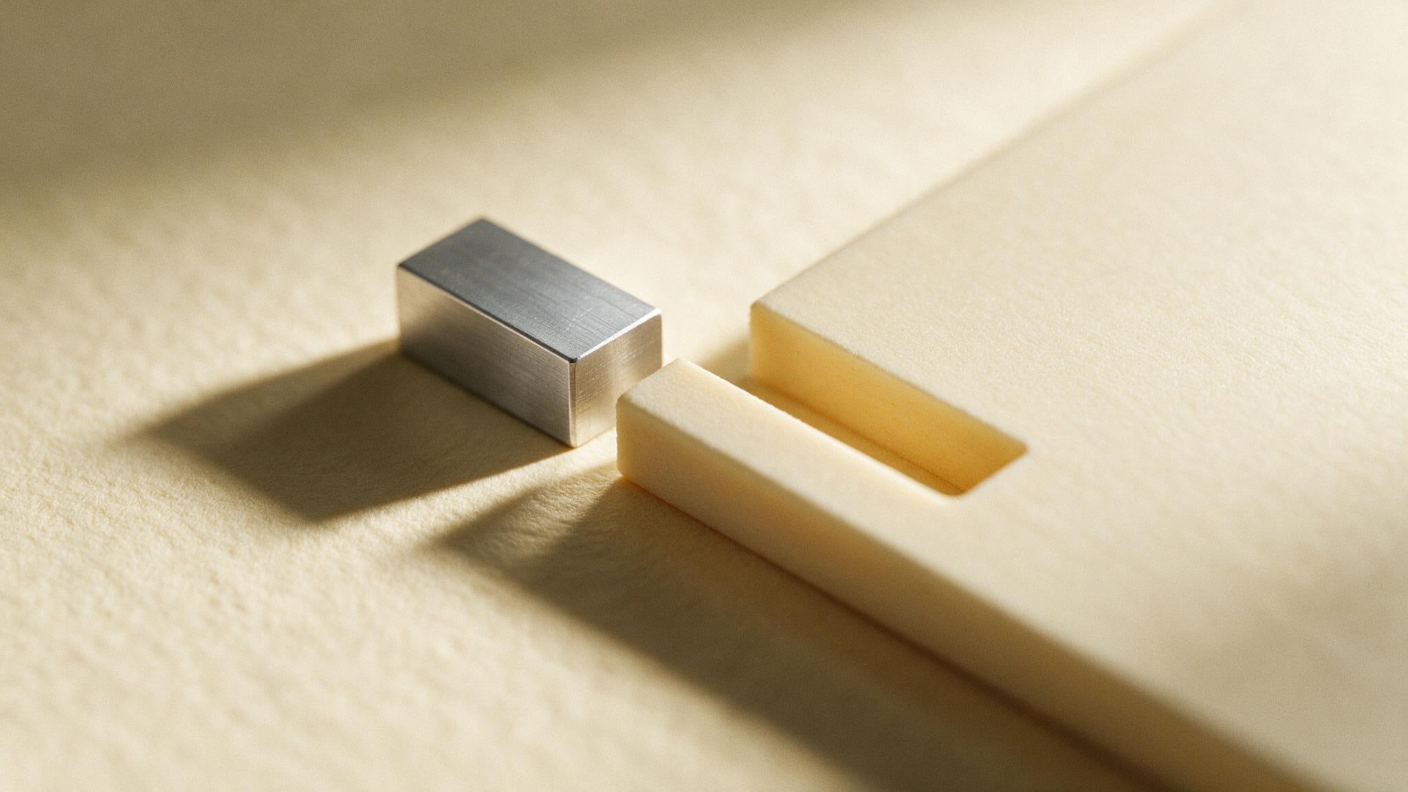 A machined aluminium block resting on warm cream paper a few millimetres from the slot cut to receive it, raking light throwing a long soft shadow between the two.