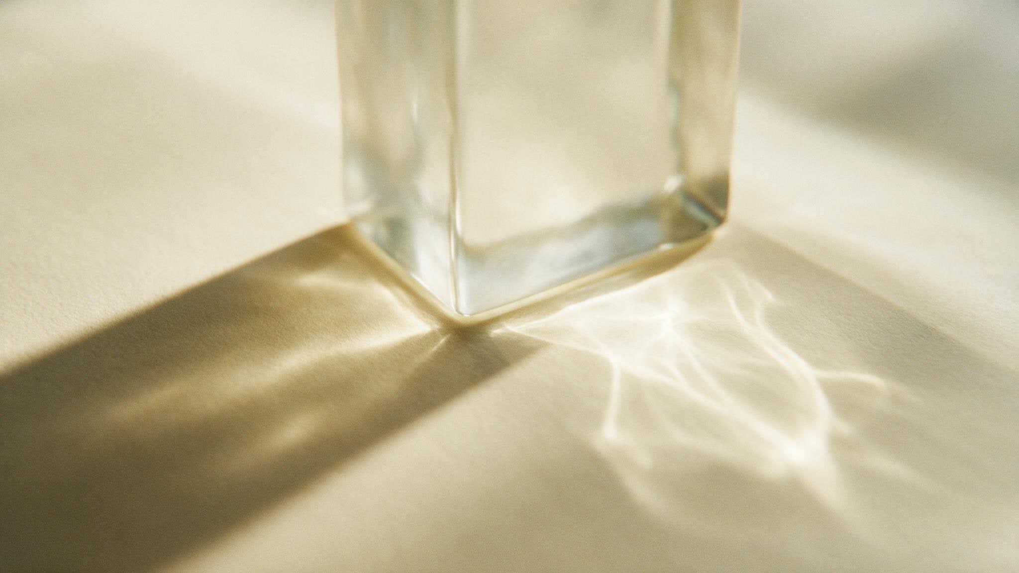 A solid glass block on warm cream paper, bending a shaft of low light into a bright caustic beside its own shadow.