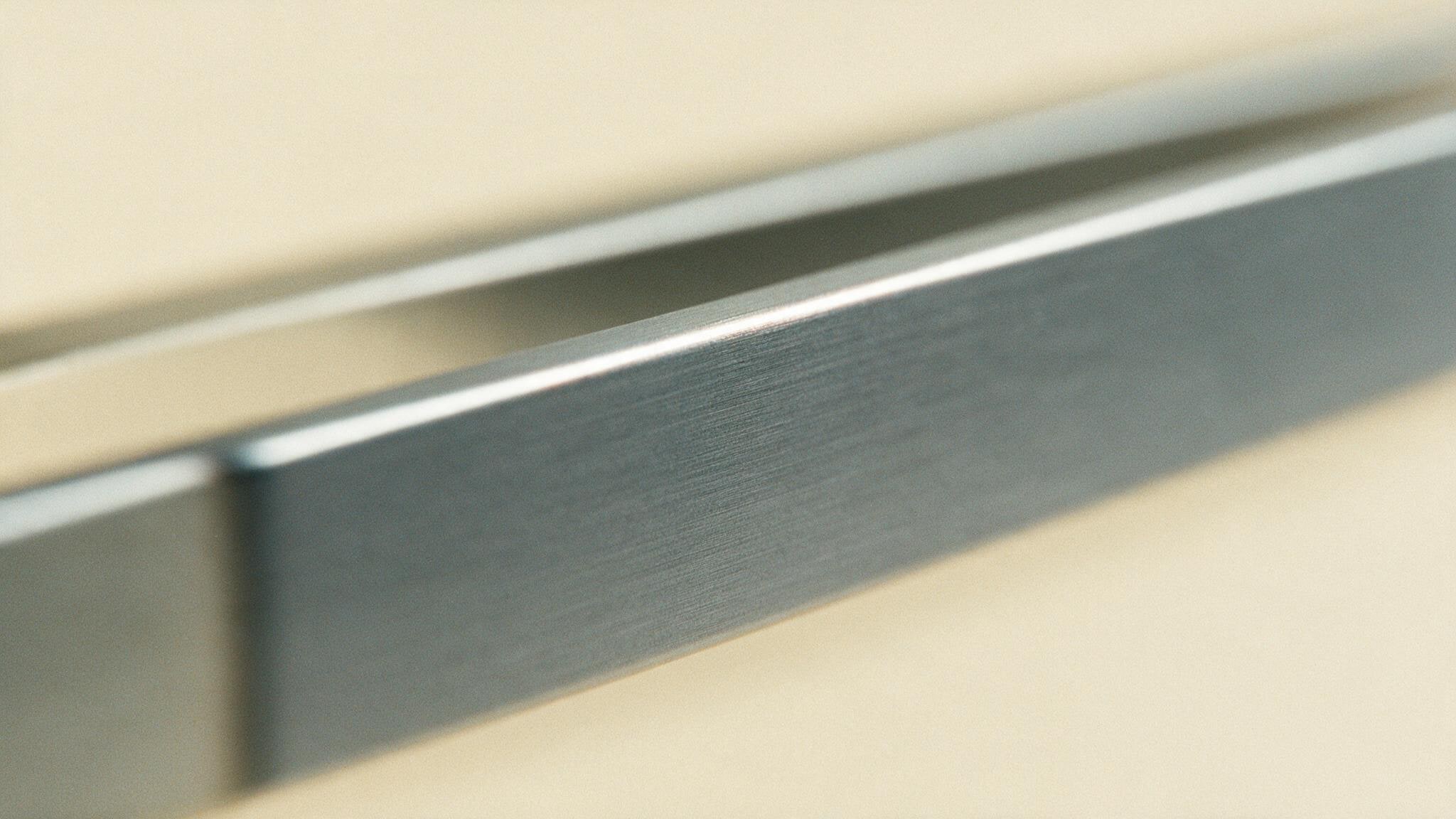 A brushed metal edge lying across warm cream, catching a single thin highlight along its bevel, a second edge receding out of focus behind it.