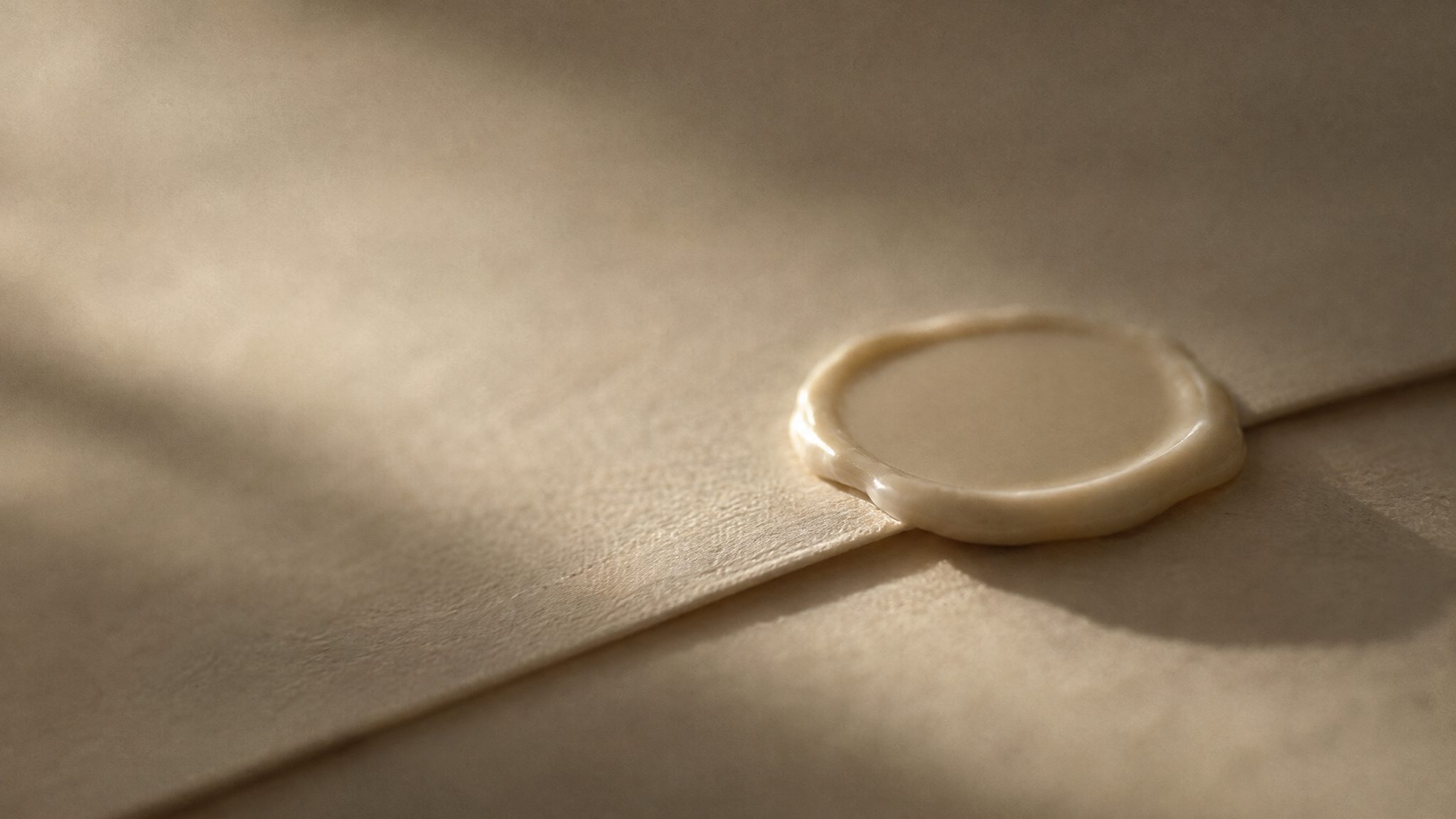 A smooth ivory disc of sealing wax, not yet stamped, closing the fold of a sheet of cream paper. Low side light picks out the poured edge of the wax and casts a long shadow.